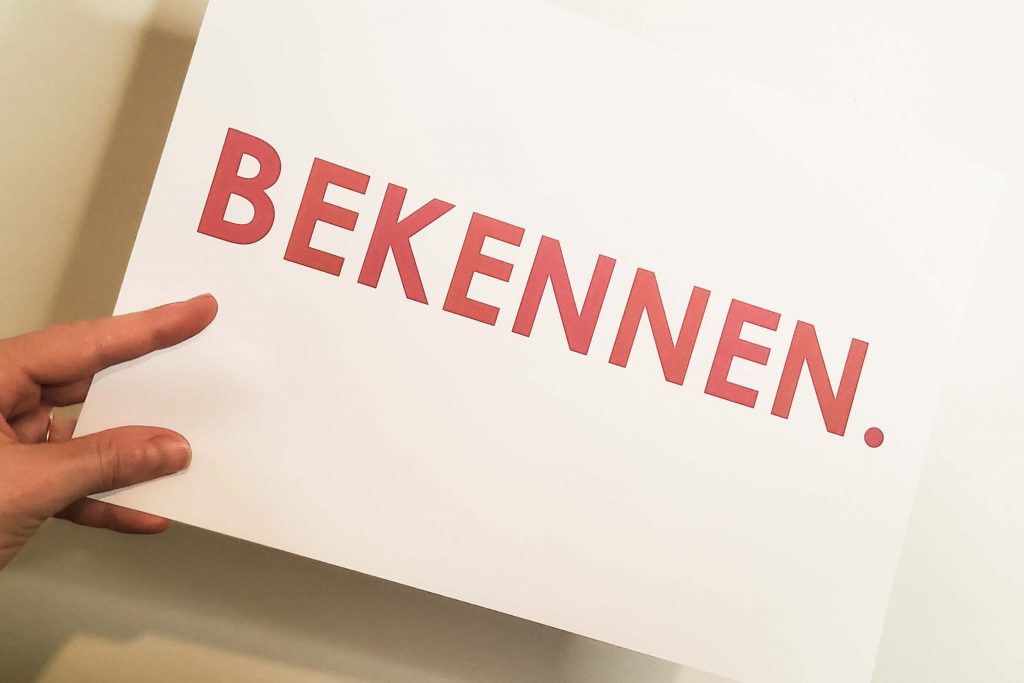 eine Hand hält ein Schild mit "Bekennen" in die Kamera