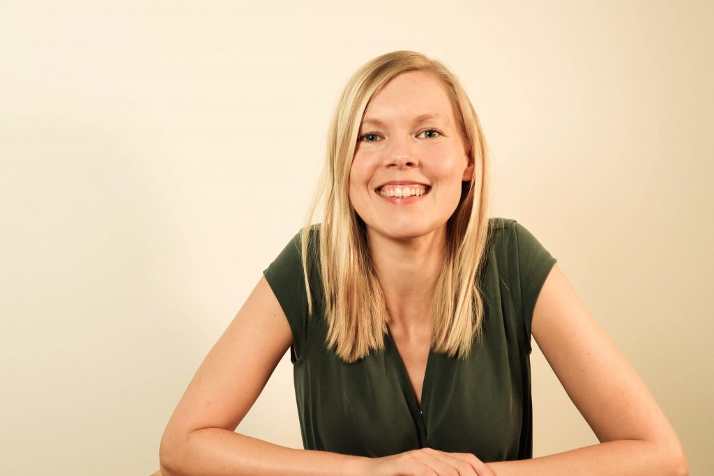 blonde Frau lacht in die Kamera und hat ihre Arme locker vor sich auf der Unterlage abgelegt